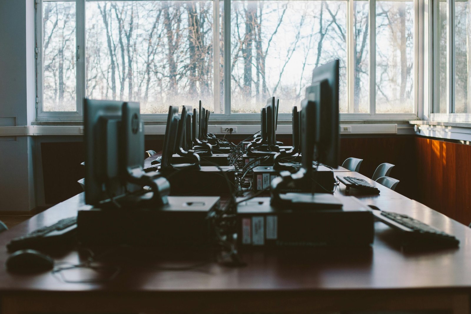 An empty computer lab with multiple workstations and large windows during the daytime.