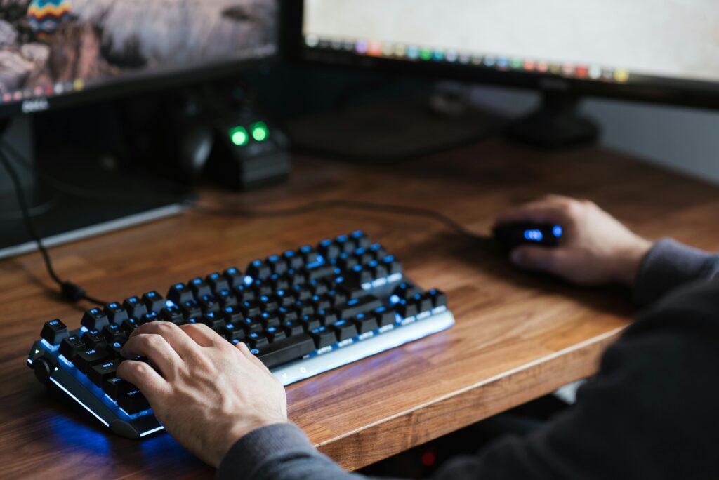 High angle crop male in casual clothes sitting at desk and browsing contemporary computer while typing on backlit keyboard and using mouse