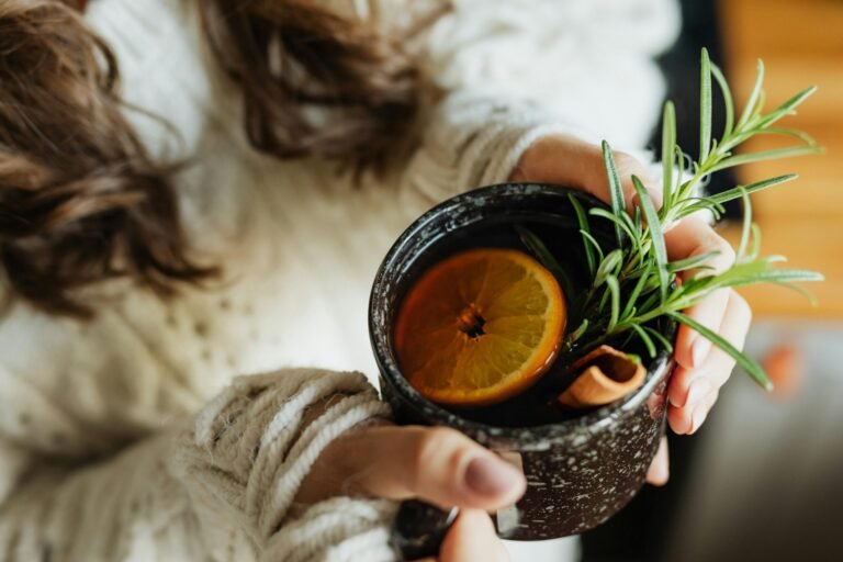 Close-up of a woman holding a mug of herbal tea with lemon and rosemary sprigs.