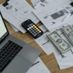 A desk with financial documents, currency, a laptop, and phone calculator.