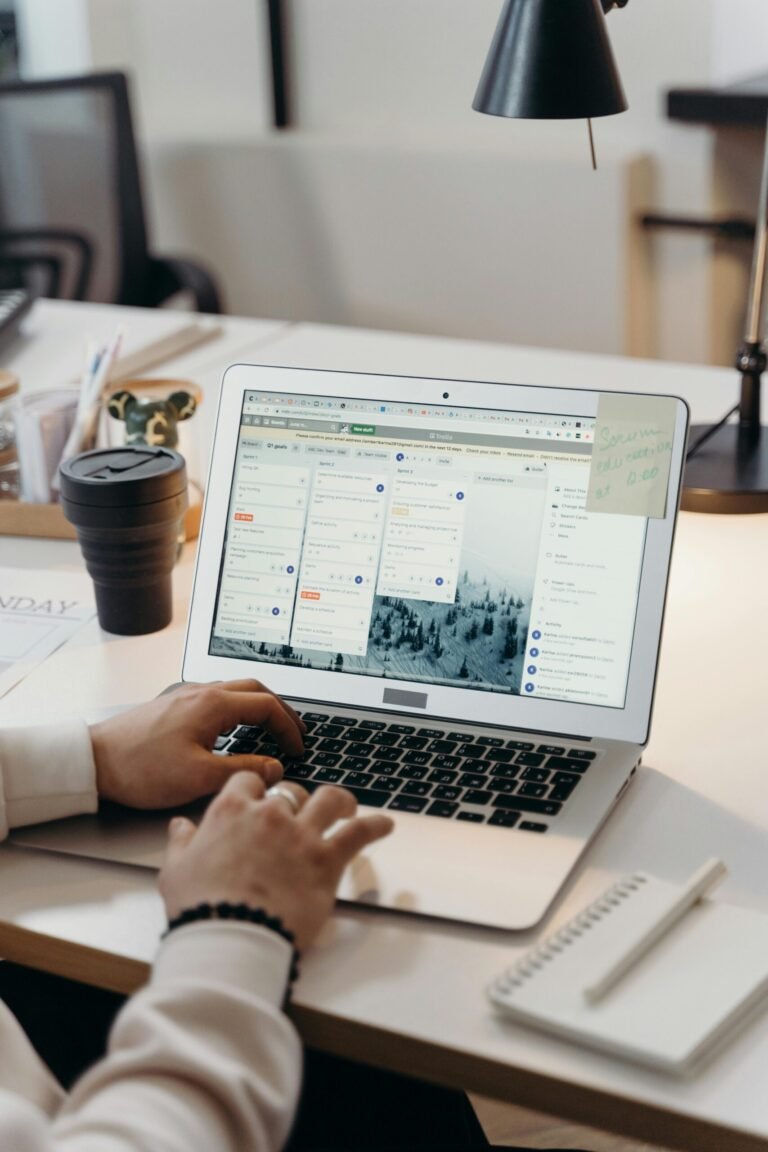 Hands typing on a laptop in a modern office setting with notes and task management software visible on the screen.