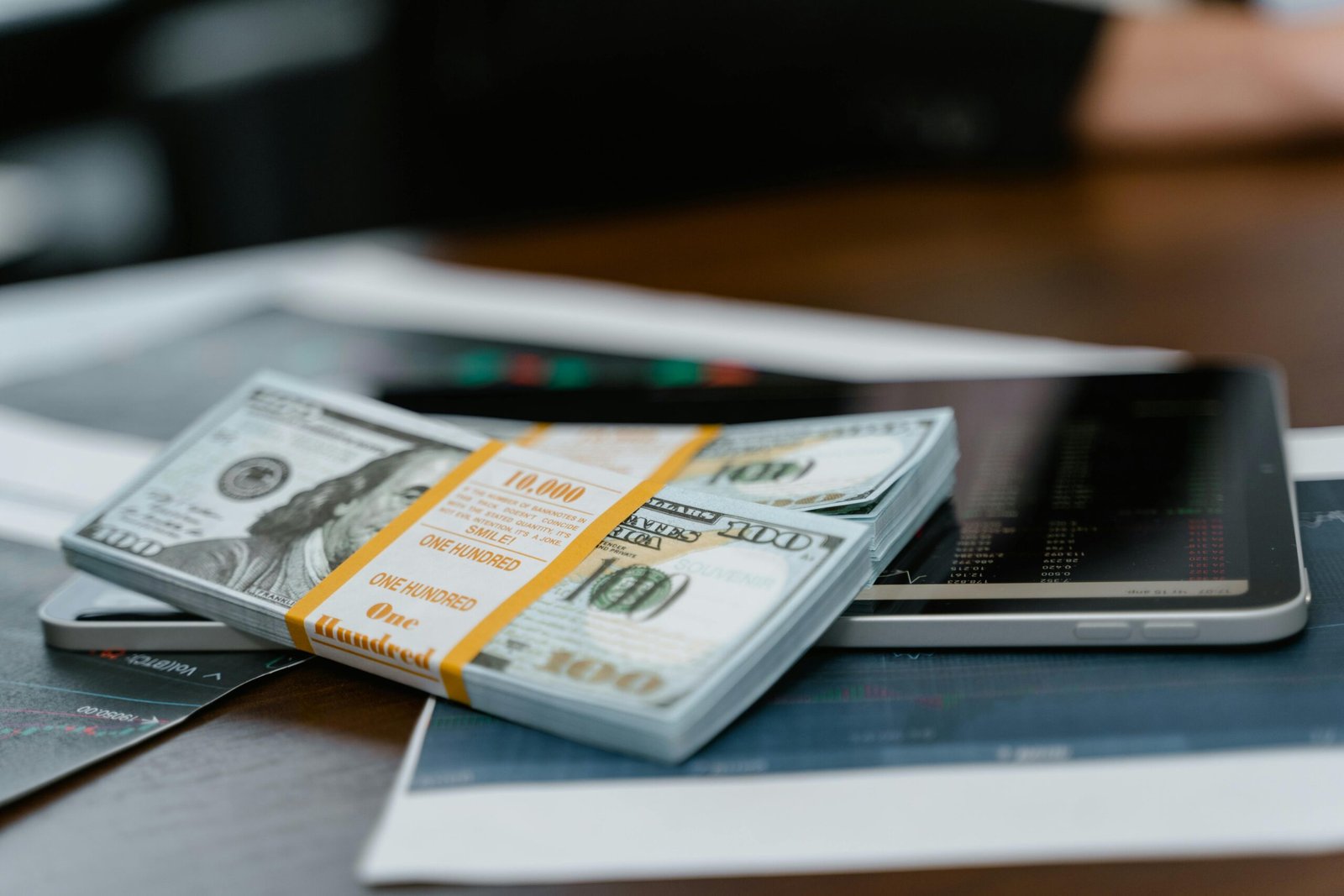 A stack of $100 bills placed on a tablet displaying financial graphs, representing modern finance.